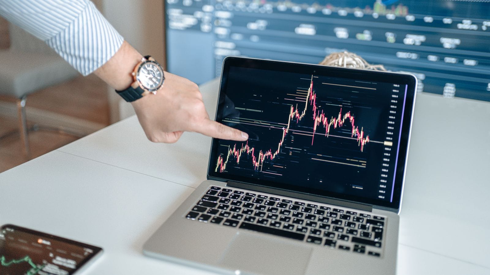 A hand pointing at a candlestick price chart on a laptop screen, on a bright desk